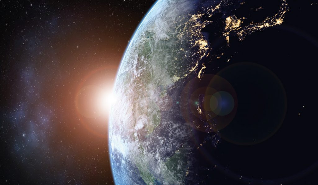 An image of planet Earth from above, depicting lights on the landmass and the sun in the background.