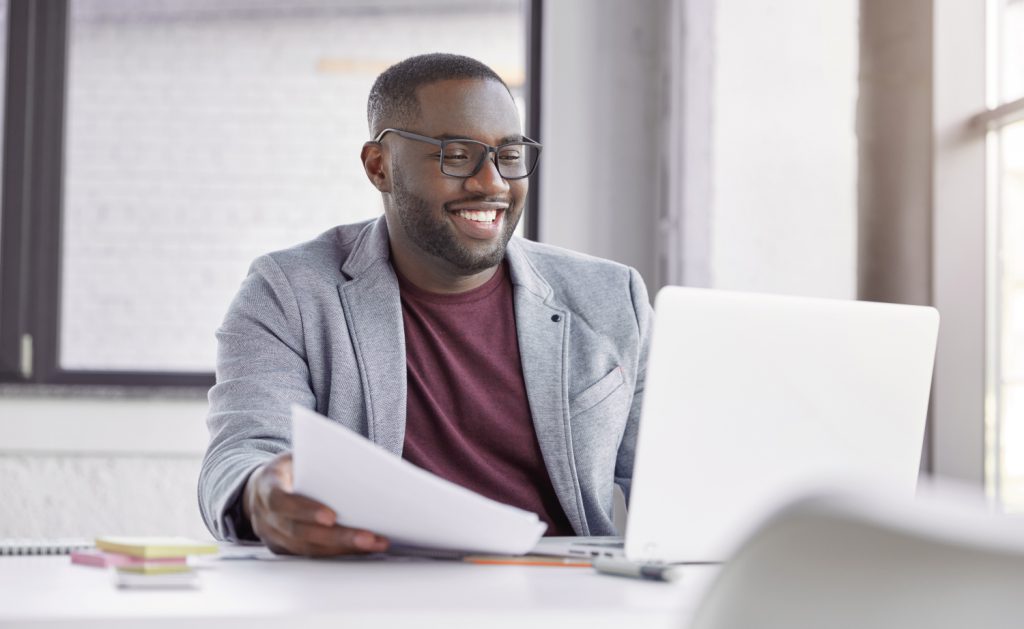 A male wearing glasses, a grey jacket and burgundy tshirt sits in front of a laptop and smiles.