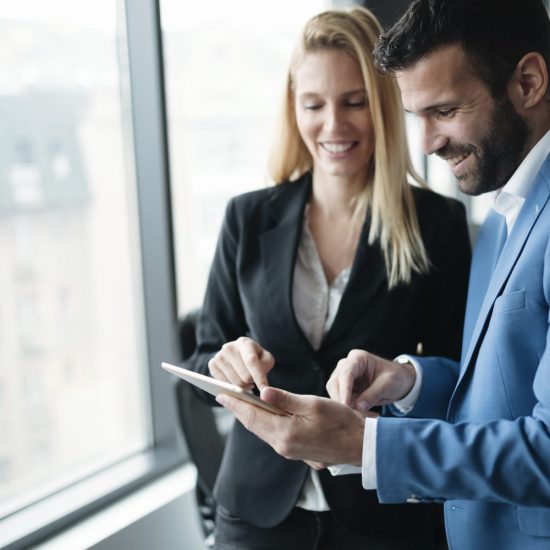 Ein Mann und eine Frau, beide im Anzug, lächeln über ein Tablet.