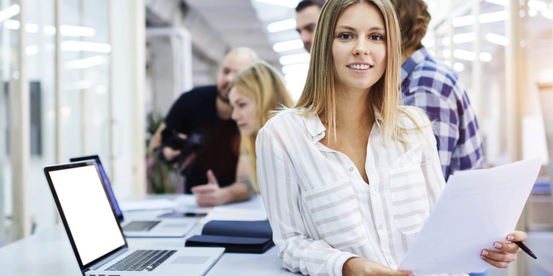 A woman leaning against a desk with a laptop on smiles at the camera as a group of people talk in the background.