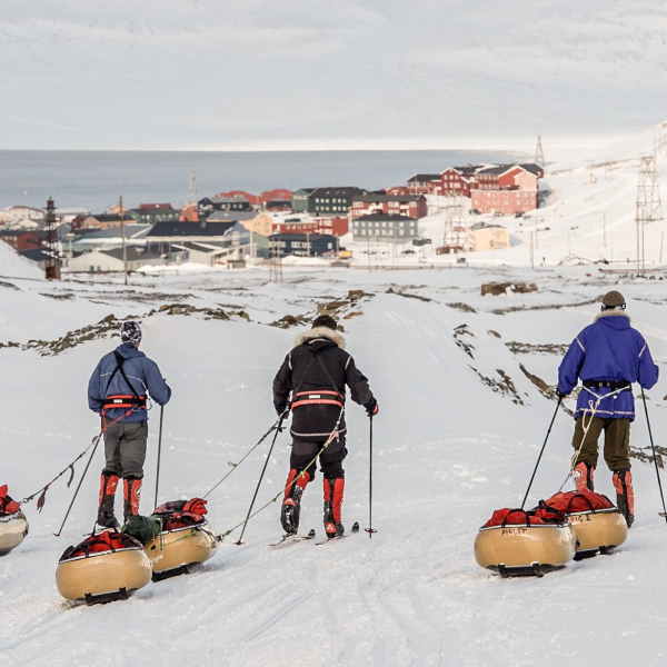 Drei Personen, die auf Skiern durch den Schnee fahren und Taschen hinter sich herziehen, fahren aus der Kamera heraus zu einem kleinen Dorf mit bunten Häusern in der Ferne.
