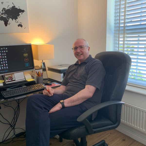 Ein Mann sitzt in einem Bürostuhl vor einem Desktop-Computer und lächelt in die Kamera.