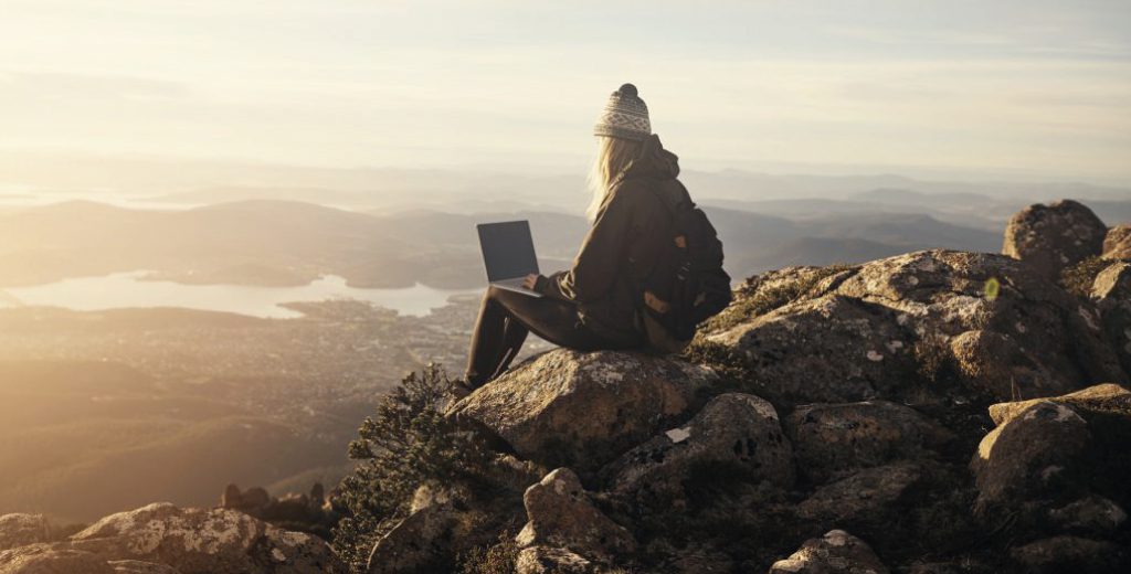 A woman wearing a bobble hat sits on rocks at the top of mountains with a laptop across her knees, looking out at the view below.