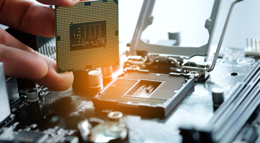 A hand holds up a computer chip above other computing hardware.