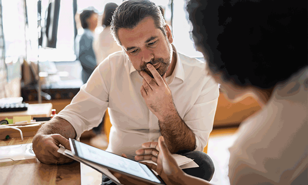 A man sitting in desk looks to be deep in thought as he looks at a tablet device held by a woman.