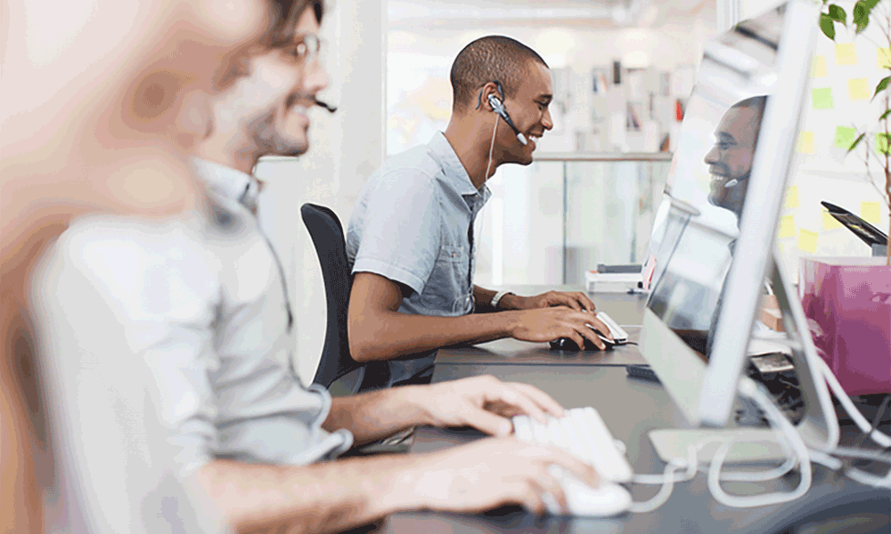 A row of three people sit in front of computers on a desk. The third person is in focus and is smiling at the screen.