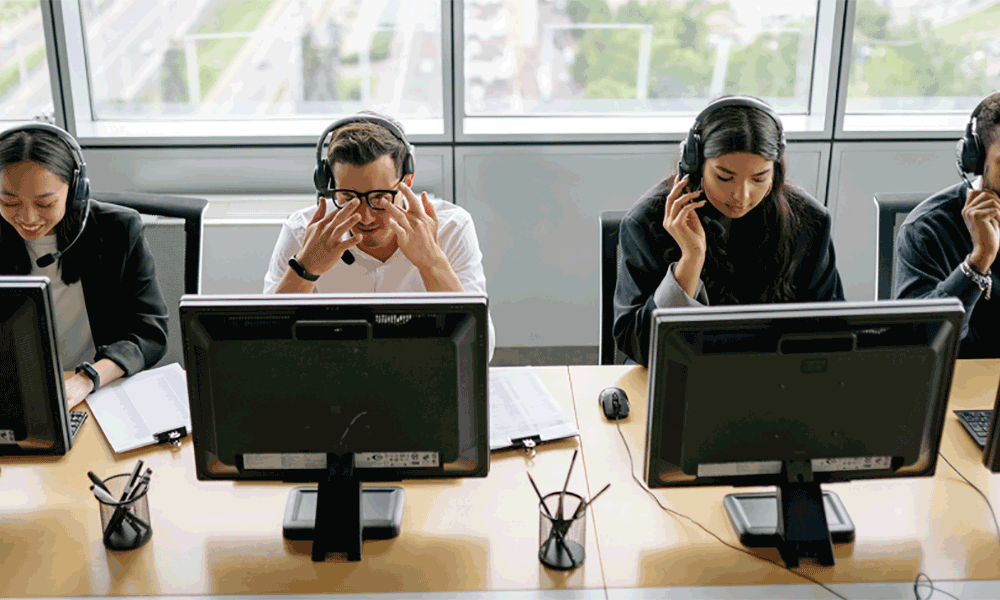A row of four people sit in front of screens on a desk wearing headsets. Some are smiling, some look exasperated.