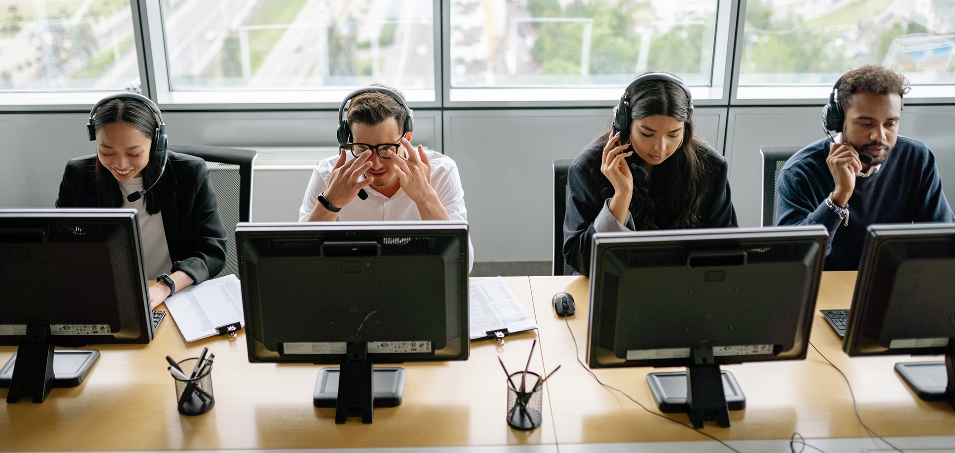 A row of four people sit in front of screens on a desk wearing headsets. Some are smiling, some look exasperated.