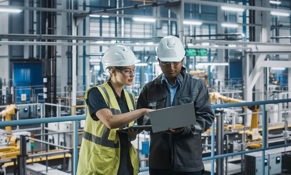 A male and female both dressed in white hard hats against an industrial backdrop look at a laptop and tablet device.